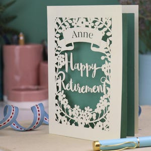 This lovely laser cut retirement card is shown standing on a white desk with a pen and ceramics in the background. It has the words Happy Retirement cut in a script font, has a banner above for the retiree's name and a floral decorative border. It is cut from cream cardstock and has a sage insert paper