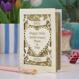A cream-coloured, laser-cut anniversary card with the text "Happy 50th Anniversary Mum & Dad" inside a heart shape. The card includes the years 1976 and 2026. A pink pen with gold accents rests in front of the card.