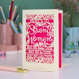K&ouml;nnte beinhalten: Eine cremefarbene, lasergeschnittene Gru&szlig;karte mit einer leuchtend pinken Einlage. Die Vorderseite zeigt florale Muster und den Text: HERE'S TO Strong Women MAY WE know THEM MAY WE be THEM MAY WE raise THEM. Die Karte steht aufrecht auf einer wei&szlig;en Fl&auml;che.