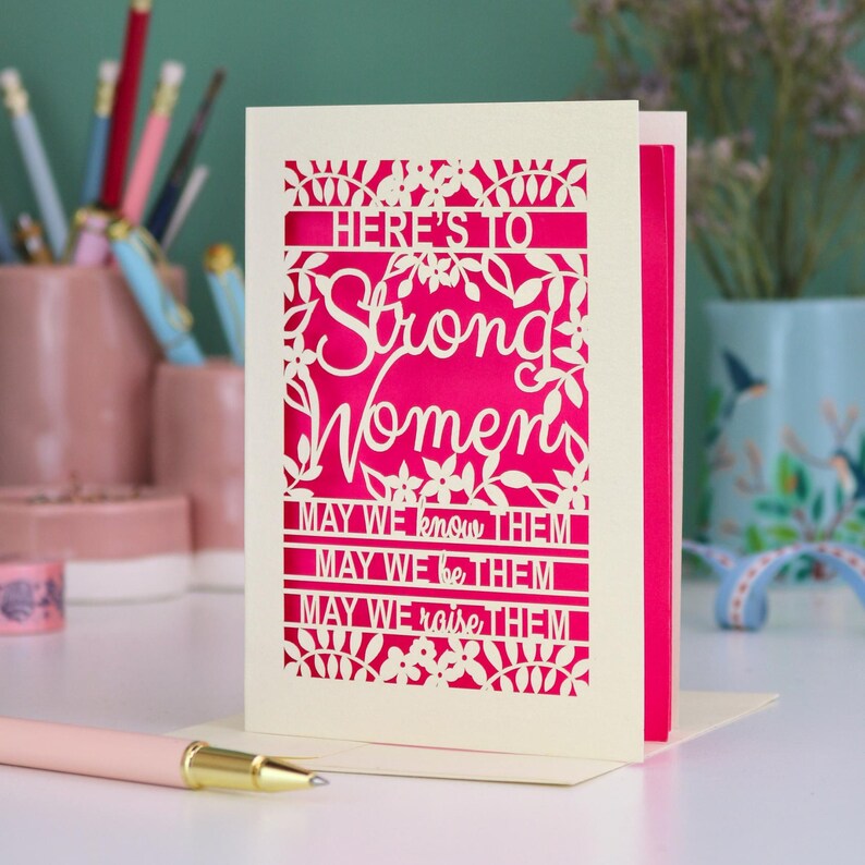 K&ouml;nnte beinhalten: Eine cremefarbene, lasergeschnittene Gru&szlig;karte mit einer leuchtend pinken Einlage. Die Vorderseite zeigt florale Muster und den Text: HERE'S TO Strong Women MAY WE know THEM MAY WE be THEM MAY WE raise THEM. Die Karte steht aufrecht auf einer wei&szlig;en Fl&auml;che.
