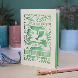 A gorgeous new baby card depicting a stork carrying a bundle surrounded with leaves, flowers and clouds. A banner saying Welcome To The World across the top,  and a second banner below with the baby's date of birth and their name under that. Shown standing on a white desk with stationery items in the fore and background. Its cut from cream cardstock and has a light green insert paper.