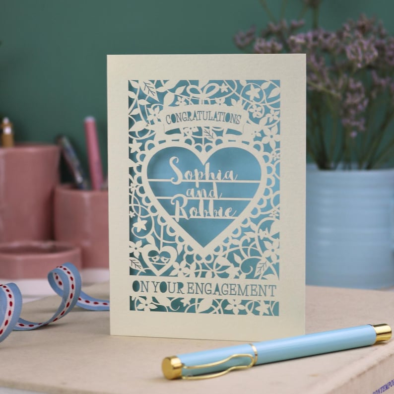 A laser cut engagement card with intricate floral cut out design, the word "Congratulations" in a banner at the top, a heart shape border with 3 lines of text, 'Name and Name', and 'On Your Engagement' in cut out lettering across the bottom of the card.