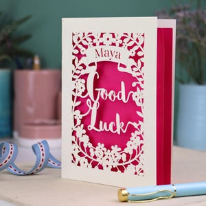 This floral Good Luck card is shown standing on a book with a pen, ribbon and ceramics in the background. It shows the text Good Luck in the centre of the card with a banner above showing the name Maya. It is laser cut from cream card with a shocking pink insert paper