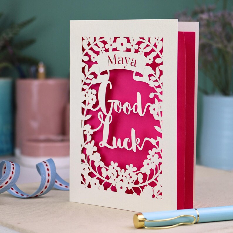 This floral Good Luck card is shown standing on a book with a pen, ribbon and ceramics in the background. It shows the text Good Luck in the centre of the card with a banner above showing the name Maya. It is laser cut from cream card with a shocking pink insert paper
