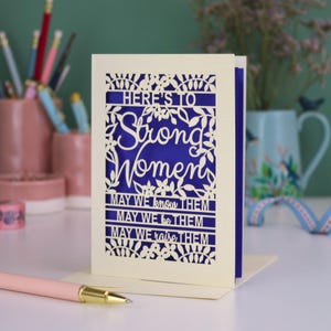 K&ouml;nnte beinhalten: Eine cremefarbene, lasergeschnittene Gru&szlig;karte mit einem tiefblauen Einleger. Die Vorderseite zeigt florale Muster und den Text: HERE'S TO Strong Women MAY WE know THEM MAY WE be THEM MAY WE raise THEM. Die Karte steht aufrecht auf einer wei&szlig;en Oberfl&auml;che.