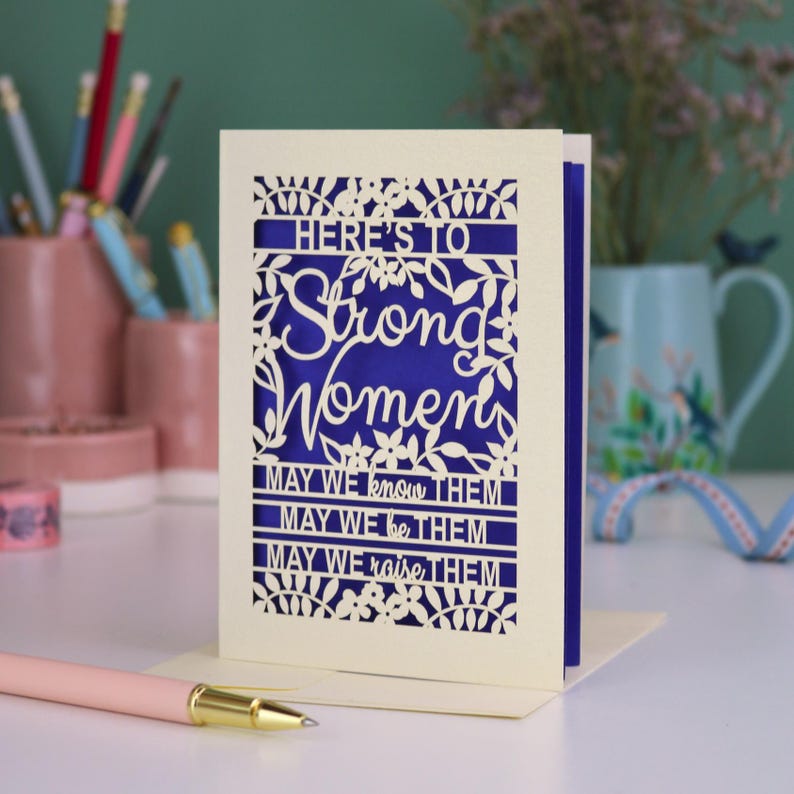 K&ouml;nnte beinhalten: Eine cremefarbene, lasergeschnittene Gru&szlig;karte mit einem tiefblauen Einleger. Die Vorderseite zeigt florale Muster und den Text: HERE'S TO Strong Women MAY WE know THEM MAY WE be THEM MAY WE raise THEM. Die Karte steht aufrecht auf einer wei&szlig;en Oberfl&auml;che.