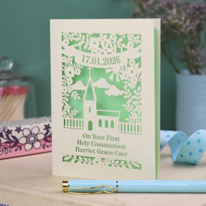 This beautiful laser cut First Holy Communion card shows a church surrounded by trees, flowers, clouds and the sun. With a banner flying above the church for the  Communion date and beneath the text "On Your First Holy Communion" and the child's name. Shown standing on a white desk with stationery items, laser cut from cream cardstock with a light green insert paper