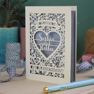 A laser cut engagement card with intricate floral cut out design, the word "Congratulations" in a banner at the top, a heart shape border with 3 lines of text, 'Name and Name', and 'On Your Engagement' in cut out lettering across the bottom of the card.