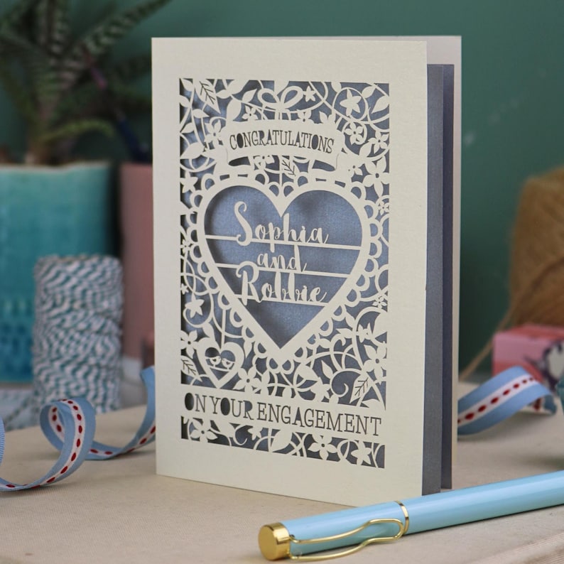 A laser cut engagement card with intricate floral cut out design, the word "Congratulations" in a banner at the top, a heart shape border with 3 lines of text, 'Name and Name', and 'On Your Engagement' in cut out lettering across the bottom of the card.