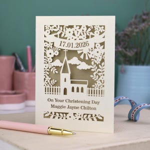 This beautiful laser cut Christening card shows a church surrounded by trees, flowers, clouds and the sun. With a banner flying above the church for the Christening date and beneath the text "On Your Christening Day" and the child's name. Shown standing on a white desk with stationery items, laser cut from cream cardstock with a gold leaf insert paper