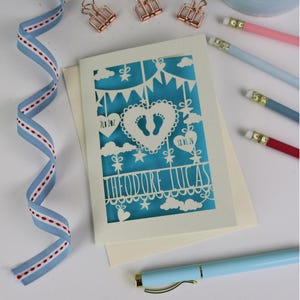 A laser cut new baby card  shown flat on a cream coloured envelope on a white desk surrounded with  stationery items and ribbon. This cute card is laser cut from a cream cardstock with a peacock blue paper insert and features a heart with a pair of baby feet cut out, bunting, clouds, stars and hearts. The text under the heart is the baby's name Theodore Lucas and the left hanging heart shows the baby's weight, 7lb 2oz, and the right hanging heart shows the baby's date of birth 18/01/26