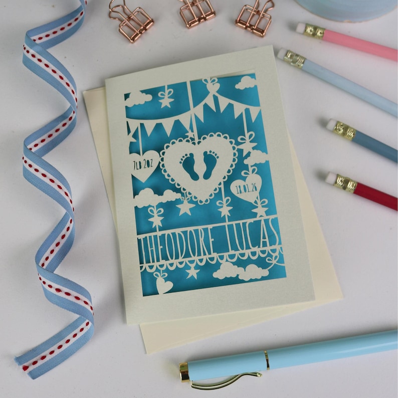 A laser cut new baby card  shown flat on a cream coloured envelope on a white desk surrounded with  stationery items and ribbon. This cute card is laser cut from a cream cardstock with a peacock blue paper insert and features a heart with a pair of baby feet cut out, bunting, clouds, stars and hearts. The text under the heart is the baby's name Theodore Lucas and the left hanging heart shows the baby's weight, 7lb 2oz, and the right hanging heart shows the baby's date of birth 18/01/26