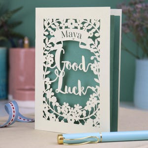 This floral Good Luck card is shown standing on a book with a pen, ribbon and ceramics in the background. It shows the text Good Luck in the centre of the card with a banner above showing the name Maya. It is laser cut from cream card with a sage insert paper