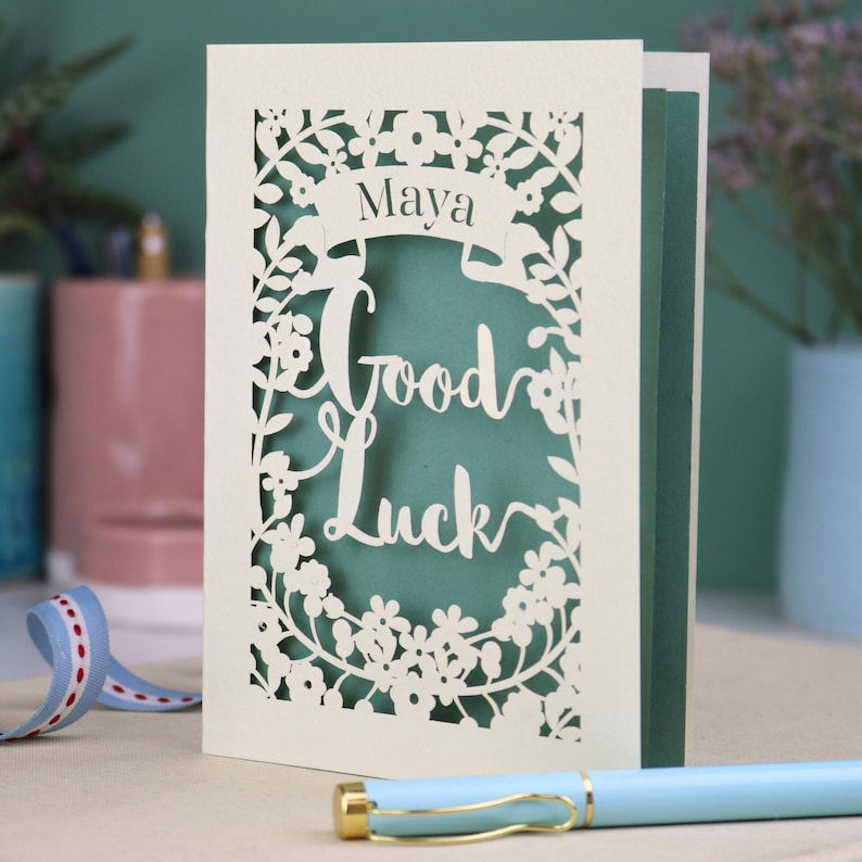 This floral Good Luck card is shown standing on a book with a pen, ribbon and ceramics in the background. It shows the text Good Luck in the centre of the card with a banner above showing the name Maya. It is laser cut from cream card with a sage insert paper