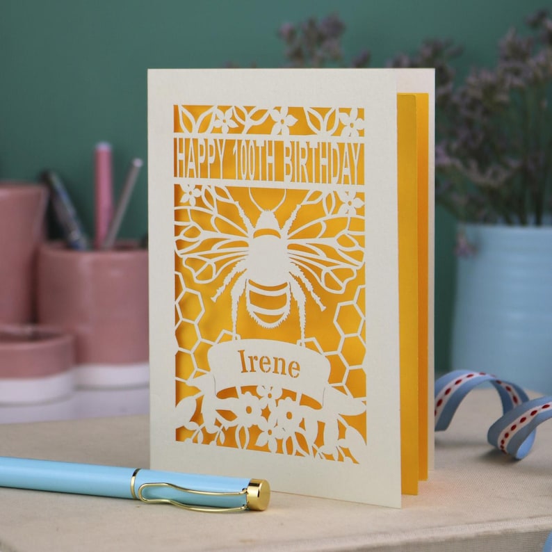 Peut inclure: Une carte de v&oelig;ux d&eacute;coup&eacute;e au laser de couleur cr&egrave;me avec un motif d'abeille et de nid d'abeille. Le texte indique HAPPY 100TH BIRTHDAY et le pr&eacute;nom Irene sur une banni&egrave;re. Une couche int&eacute;rieure jaune vif est visible &agrave; travers les d&eacute;coupes.