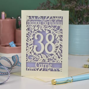 A laser cut flower birthday card with cut out floral design and "Happy Birthday" text. The birthday card is personalised with a cut out name and the age in the centre, surrounded by leaves and flowers