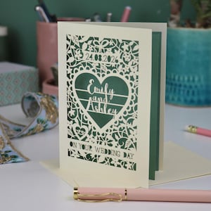 A laser cut "on your wedding day" card with floral design and cut out personalised text. Card features the wedding date, the first names of the couple in a heart shape, and the text "On your wedding day" Standing on a white desk with stationery items in the background. Cut from cream card stock with a sage insert paper