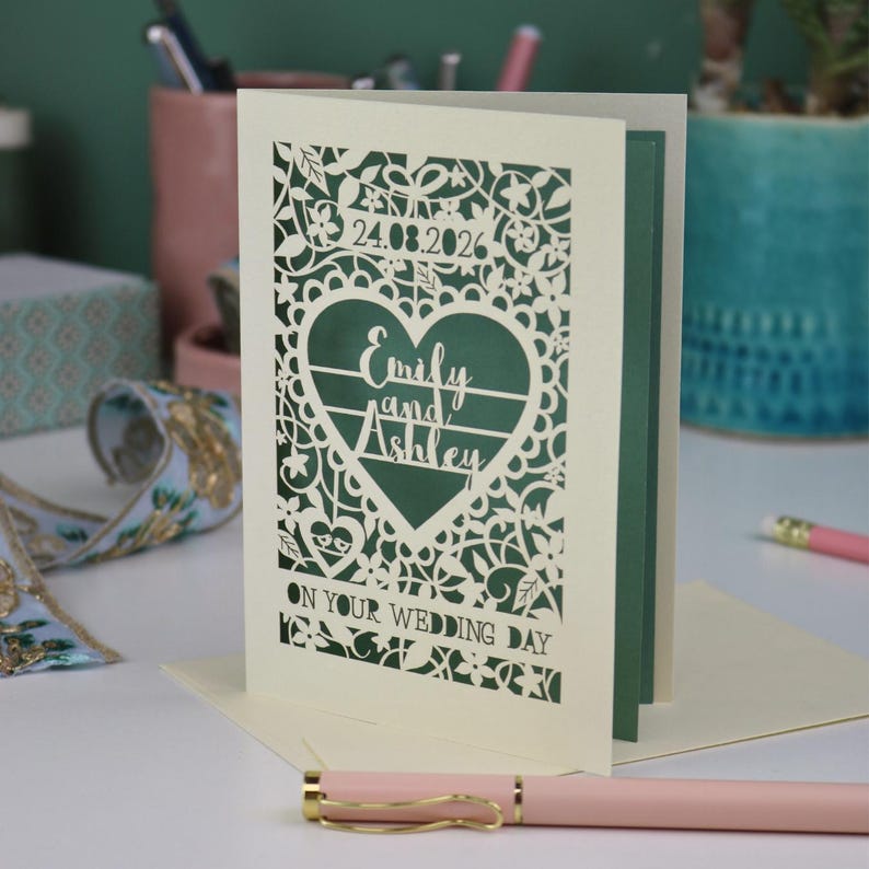 A laser cut "on your wedding day" card with floral design and cut out personalised text. Card features the wedding date, the first names of the couple in a heart shape, and the text "On your wedding day" Standing on a white desk with stationery items in the background. Cut from cream card stock with a sage insert paper