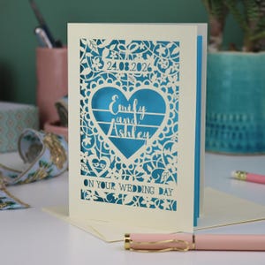 A laser cut "on your wedding day" card with floral design and cut out personalised text. Card features the wedding date, the first names of the couple in a heart shape, and the text "On your wedding day" Standing on a white desk with stationery items in the background. Cut from cream card stock with a peacock blue insert paper