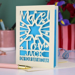 K&ouml;nnte beinhalten: Eine cremefarbene, lasergeschnittene Gru&szlig;karte mit einem Davidstern und dem Text Mazel Tov! JACK ON YOUR BAR MITZVAH. Die Karte hat eine leuchtend blaue Innenschicht, die durch die filigranen Ausstanzungen von Sternen und Strahlen sichtbar ist.