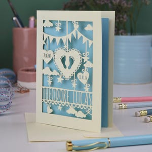 A laser cut new baby card on a white desk with stationery items in the 
background. This lovely card is laser cut from a cream cardstock with a light blue paper insert and features a heart with a pair of baby feet cut out, bunting, clouds, stars and hearts. The text under the heart is the baby's name Theodore Lucas and the left hanging heart shows the baby's weight, 7lb 2oz, and the right hanging heart shows the baby's date of birth 18/01/26
