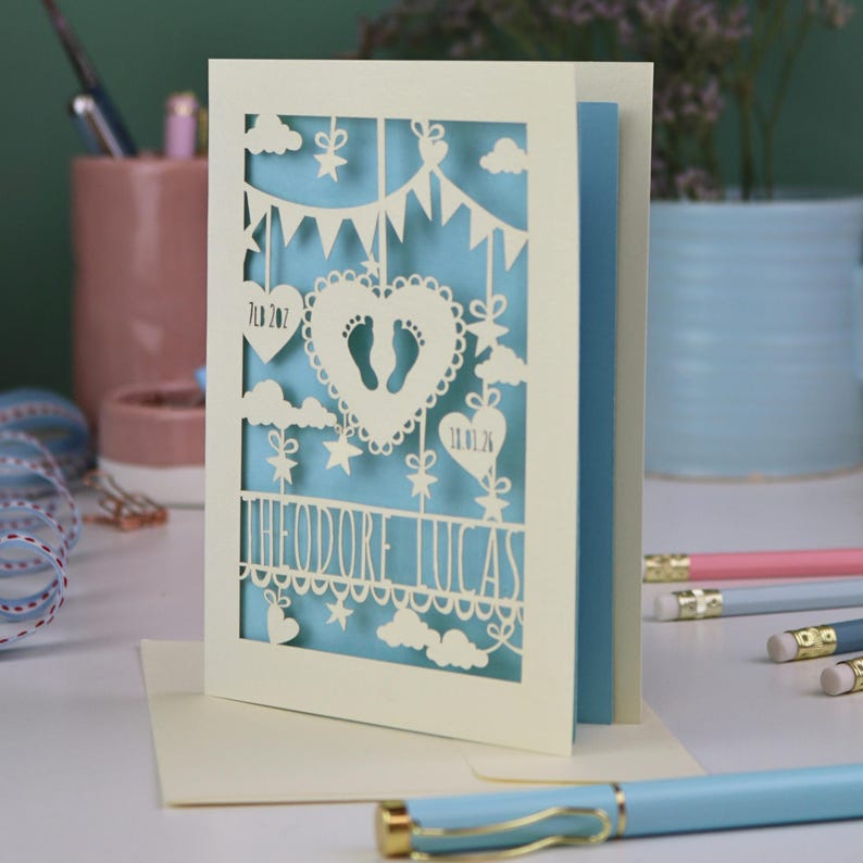 A laser cut new baby card on a white desk with stationery items in the 
background. This lovely card is laser cut from a cream cardstock with a light blue paper insert and features a heart with a pair of baby feet cut out, bunting, clouds, stars and hearts. The text under the heart is the baby's name Theodore Lucas and the left hanging heart shows the baby's weight, 7lb 2oz, and the right hanging heart shows the baby's date of birth 18/01/26