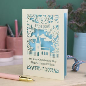 This beautiful laser cut Christening card shows a church surrounded by trees, flowers, clouds and the sun. With a banner flying above the church for the Christening date and beneath the text "On Your Christening Day" and the child's name. Shown standing on a white desk with stationery items, laser cut from cream cardstock with a light blue insert paper