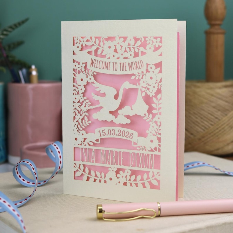 A gorgeous new baby card depicting a stork carrying a bundle surrounded with leaves, flowers and clouds. A banner saying Welcome To The World across the top,  and a second banner below with the baby's date of birth and their name under that. Shown standing on a white desk with stationery items in the fore and background. Its cut from cream cardstock and has a candy pink insert paper.