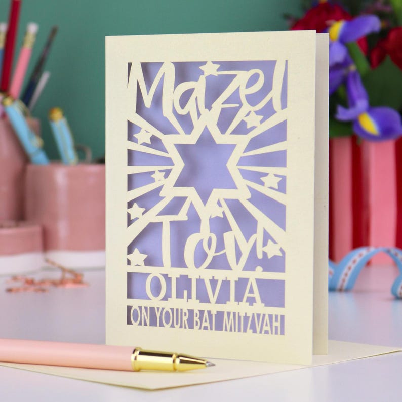 May include: A cream-coloured laser-cut greeting card featuring a Star of David, the text Mazel Tov, the name Olivia, and the phrase On Your Bat Mitzvah. The card has a light purple paper insert visible through the intricate cutouts and star shapes.