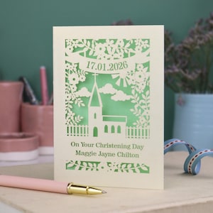 This beautiful laser cut Christening card shows a church surrounded by trees, flowers, clouds and the sun. With a banner flying above the church for the Christening date and beneath the text "On Your Christening Day" and the child's name. Shown standing on a white desk with stationery items, laser cut from cream cardstock with a light green insert paper