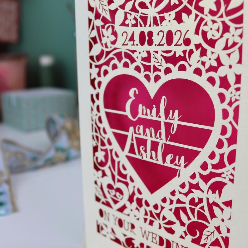 A laser cut "on your wedding day" card with floral design and cut out personalised text. Card features the wedding date, the first names of the couple in a heart shape, and the text "On your wedding day" Standing on a white desk with stationery items in the background. Cut from cream card stock with a shocking pink insert paper
