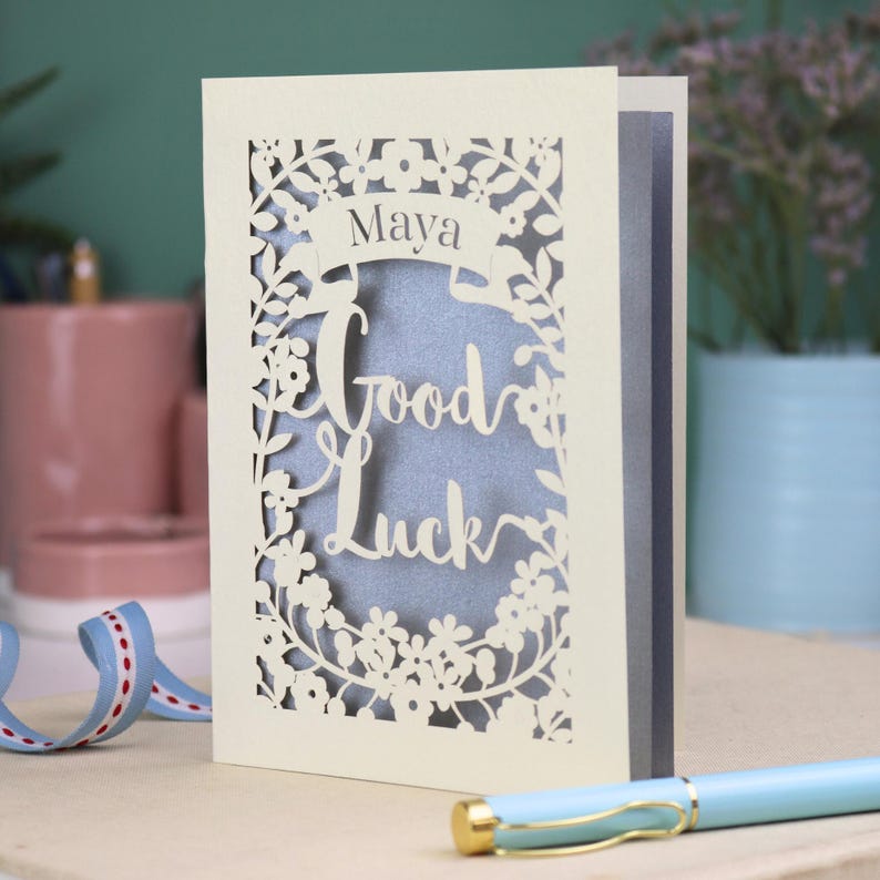 This floral Good Luck card is shown standing on a book with a pen, ribbon and ceramics in the background. It shows the text Good Luck in the centre of the card with a banner above showing the name Maya. It is laser cut from cream card with a silver insert paper