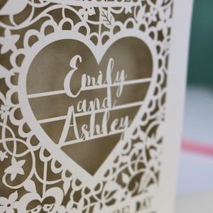 A close up detail of a laser cut "on your wedding day" card with floral design and cut out personalised text. Card features the wedding date, the first names of the couple in a heart shape, and the text "On your wedding day" Standing on a white desk with stationery items in the background. Cut from cream card stock with a gold leaf insert paper