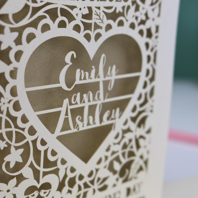 A close up detail of a laser cut "on your wedding day" card with floral design and cut out personalised text. Card features the wedding date, the first names of the couple in a heart shape, and the text "On your wedding day" Standing on a white desk with stationery items in the background. Cut from cream card stock with a gold leaf insert paper
