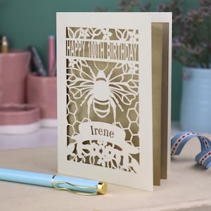Peut inclure: Une carte de v&oelig;ux d&eacute;coup&eacute;e au laser de couleur cr&egrave;me avec un motif d'abeille et de nid d'abeille. Le texte indique HAPPY 100TH BIRTHDAY et le pr&eacute;nom Irene sur une banni&egrave;re. Un panneau int&eacute;rieur dor&eacute; est visible &agrave; travers les d&eacute;coupes.