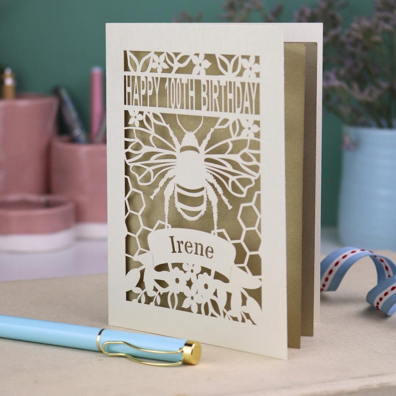 Peut inclure: Une carte de v&oelig;ux d&eacute;coup&eacute;e au laser de couleur cr&egrave;me avec un motif d'abeille et de nid d'abeille. Le texte indique HAPPY 100TH BIRTHDAY et le pr&eacute;nom Irene sur une banni&egrave;re. Un panneau int&eacute;rieur dor&eacute; est visible &agrave; travers les d&eacute;coupes.