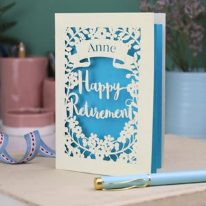 A laser cut retirement card standing on a white desk with a pen and ceramics in the background. It has the words Happy Retirement cut in a script font, has a banner above for the retiree's name and a floral decorative border. It is cut from cream cardstock and has a peacock blue insert paper
