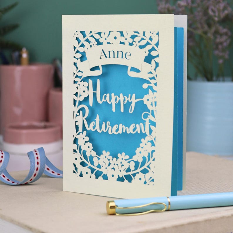 A laser cut retirement card standing on a white desk with a pen and ceramics in the background. It has the words Happy Retirement cut in a script font, has a banner above for the retiree's name and a floral decorative border. It is cut from cream cardstock and has a peacock blue insert paper