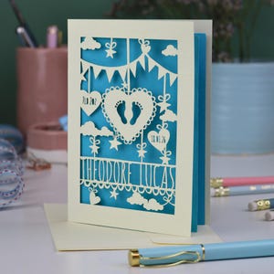 A laser cut new baby card on a white desk with stationery items in the 
background. This cute card is laser cut from a cream cardstock with a peacock blue paper insert and features a heart with a pair of baby feet cut out, bunting, clouds, stars and hearts. The text under the heart is the baby's name Theodore Lucas and the left hanging heart shows the baby's weight, 7lb 2oz, and the right hanging heart shows the baby's date of birth 18/01/26