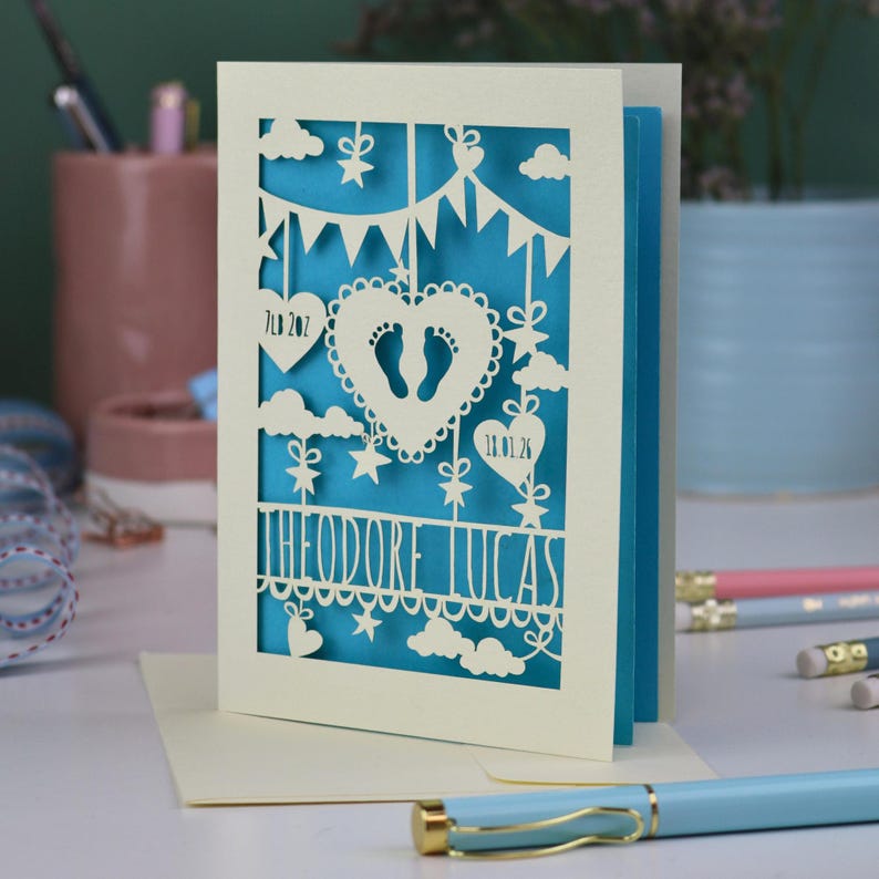 A laser cut new baby card on a white desk with stationery items in the 
background. This cute card is laser cut from a cream cardstock with a peacock blue paper insert and features a heart with a pair of baby feet cut out, bunting, clouds, stars and hearts. The text under the heart is the baby's name Theodore Lucas and the left hanging heart shows the baby's weight, 7lb 2oz, and the right hanging heart shows the baby's date of birth 18/01/26