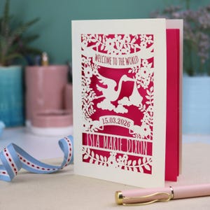 A gorgeous new baby card depicting a stork carrying a bundle surrounded with leaves, flowers and clouds. A banner saying Welcome To The World across the top,  and a second banner below with the baby's date of birth and their name under that. Shown standing on a white desk with stationery items in the fore and background. Its cut from cream cardstock and has a shocking pink insert paper.
