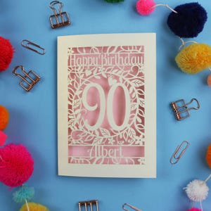 A laser cut keepsake 90th birthday card, personalised with a name in the bottom line of text. Card says Happy Birthday, 90. There is a floral border. Card is cream with a candy pink insert