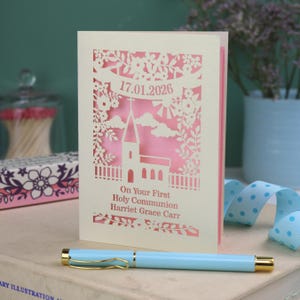 This beautiful laser cut First Holy Communion card shows a church surrounded by trees, flowers, clouds and the sun. With a banner flying above the church for the  Communion date and beneath the text "On Your First Holy Communion" and the child's name. Shown standing on a white desk with stationery items, laser cut from cream cardstock with a candy pink insert paper