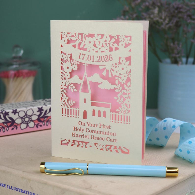 This beautiful laser cut First Holy Communion card shows a church surrounded by trees, flowers, clouds and the sun. With a banner flying above the church for the  Communion date and beneath the text "On Your First Holy Communion" and the child's name. Shown standing on a white desk with stationery items, laser cut from cream cardstock with a candy pink insert paper