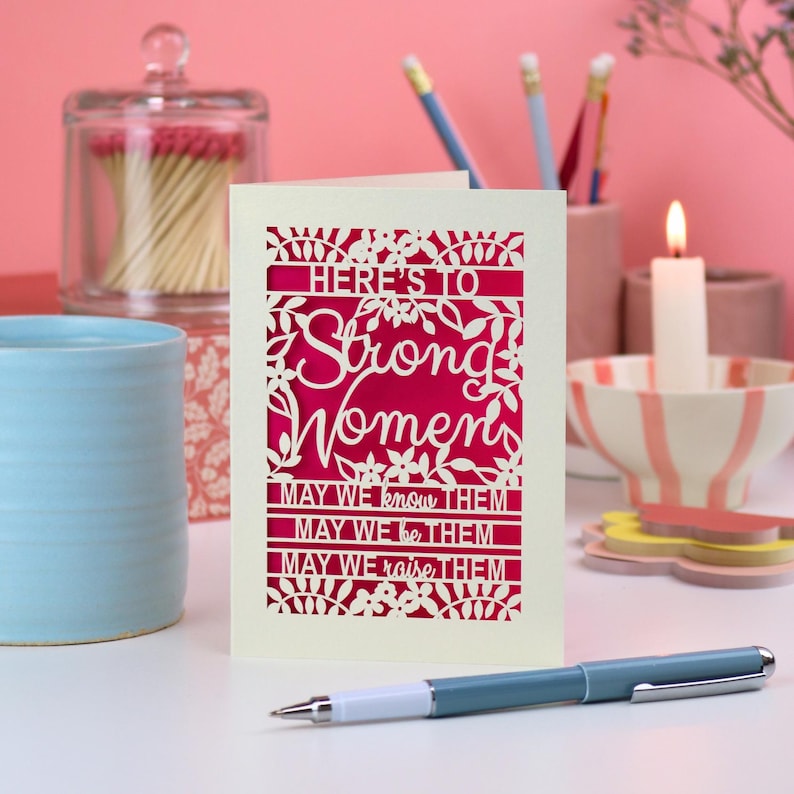 K&ouml;nnte beinhalten: Eine cremefarbene Gru&szlig;karte mit einem lasergeschnittenen Blumenrand und dem Text: "HERE'S TO Strong Women MAY WE know THEM MAY WE be THEM MAY WE raise THEM." Der Ausschnitt gibt einen leuchtend magentafarbenen Hintergrund frei.