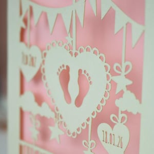 A close up view of the details on a laser cut new baby card  This cute card is laser cut from a cream cardstock with a candy pink paper insert and features a heart with a pair of baby feet cut out, bunting, clouds, stars and hearts. The text under the heart is the baby's name Theodore Lucas and the left hanging heart shows the baby's weight, 7lb 2oz, and the right hanging heart shows the baby's date of birth 18/01/26