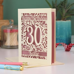 A laser cut 30th birthday card, with "Happy Birthday" text at the top, the number 30 in the centre with a floral and leaf cut out border, and personalised with a name underneath. Card is cream with a dusky pink paper insert.