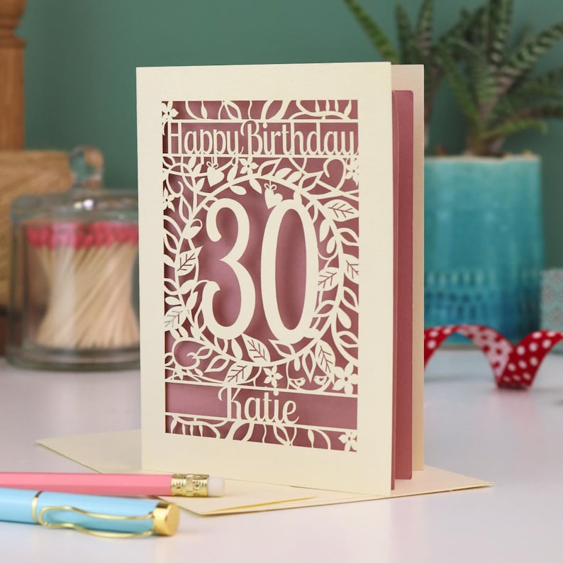A laser cut 30th birthday card, with "Happy Birthday" text at the top, the number 30 in the centre with a floral and leaf cut out border, and personalised with a name underneath. Card is cream with a dusky pink paper insert.