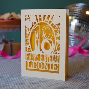 May include: A cream-coloured laser-cut birthday card featuring the number 18, star and balloon motifs, and the text HAPPY BIRTHDAY LEONIE. The card has a solid cream front layer with intricate cutouts revealing a bright yellow inner insert.