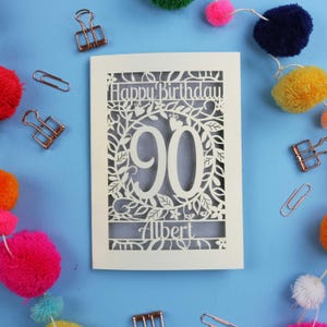 A laser cut keepsake 90th birthday card, personalised with a name in the bottom line of text. Card says Happy Birthday, 90. There is a floral border. Card is cream with a silver insert